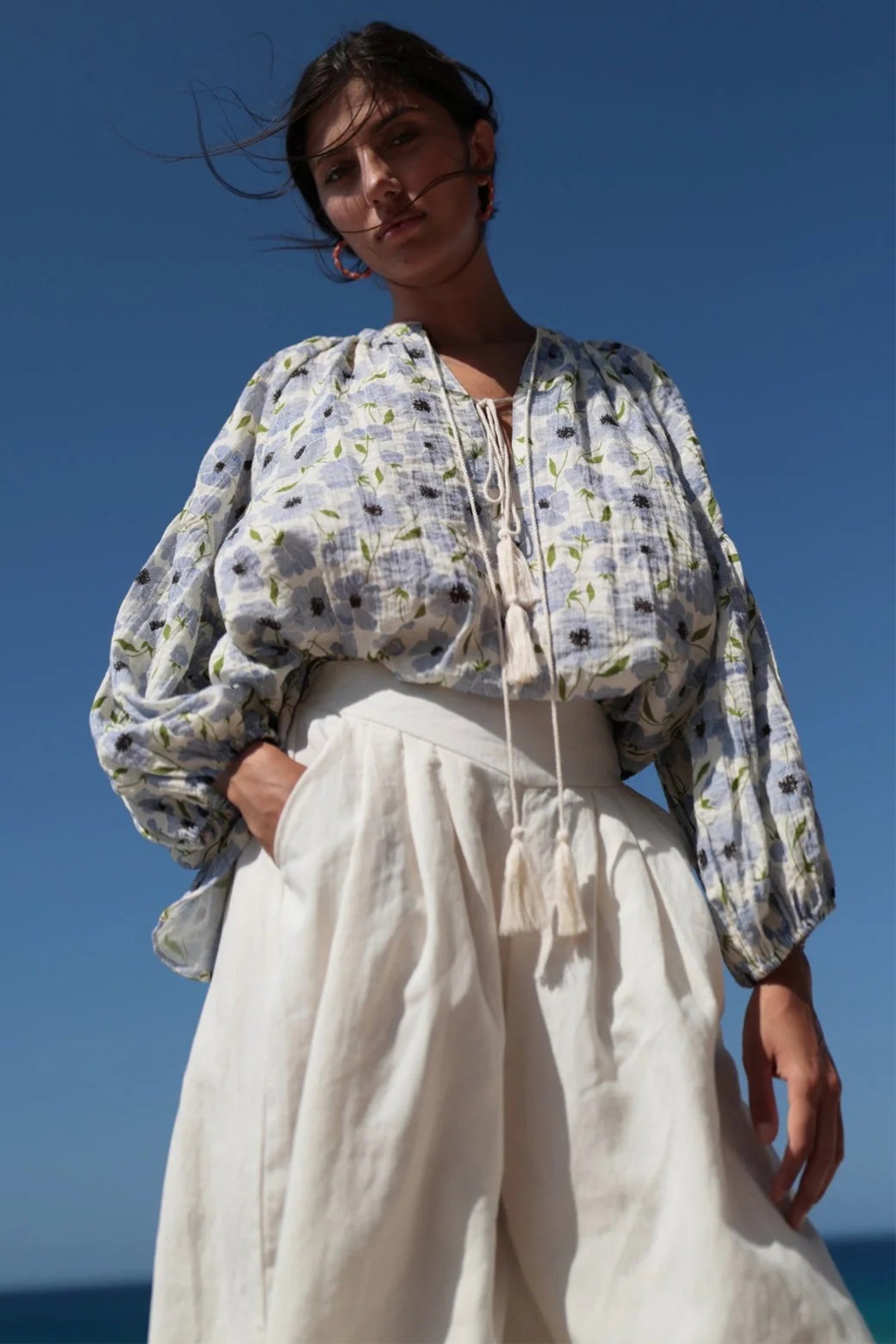 Woman wearing a floral blouse and white pants against a clear blue sky.