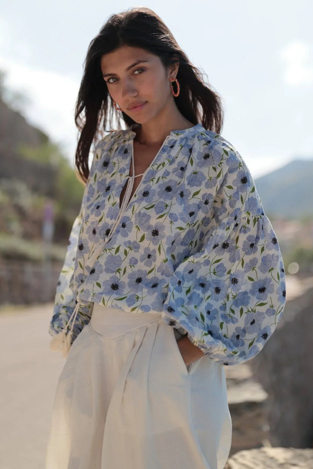 Woman wearing a floral blouse and white pants outdoors with mountains in the background