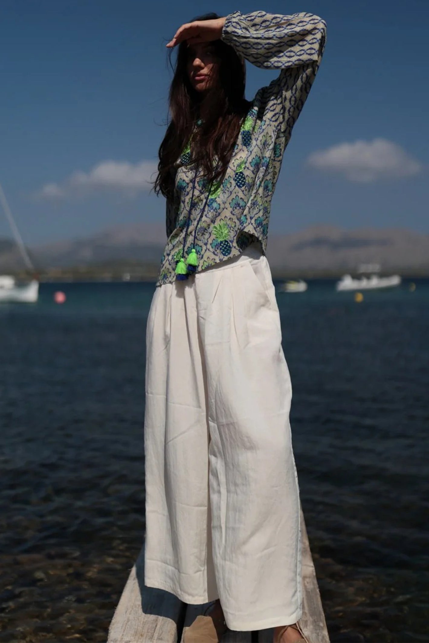 Woman standing by a body of water wearing a patterned top and white pants.