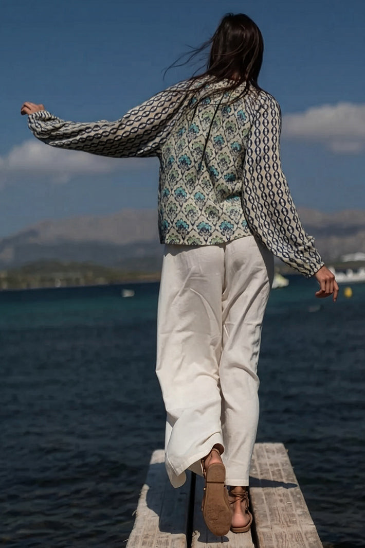 Person standing on a dock by water wearing a patterned jacket and white pants.