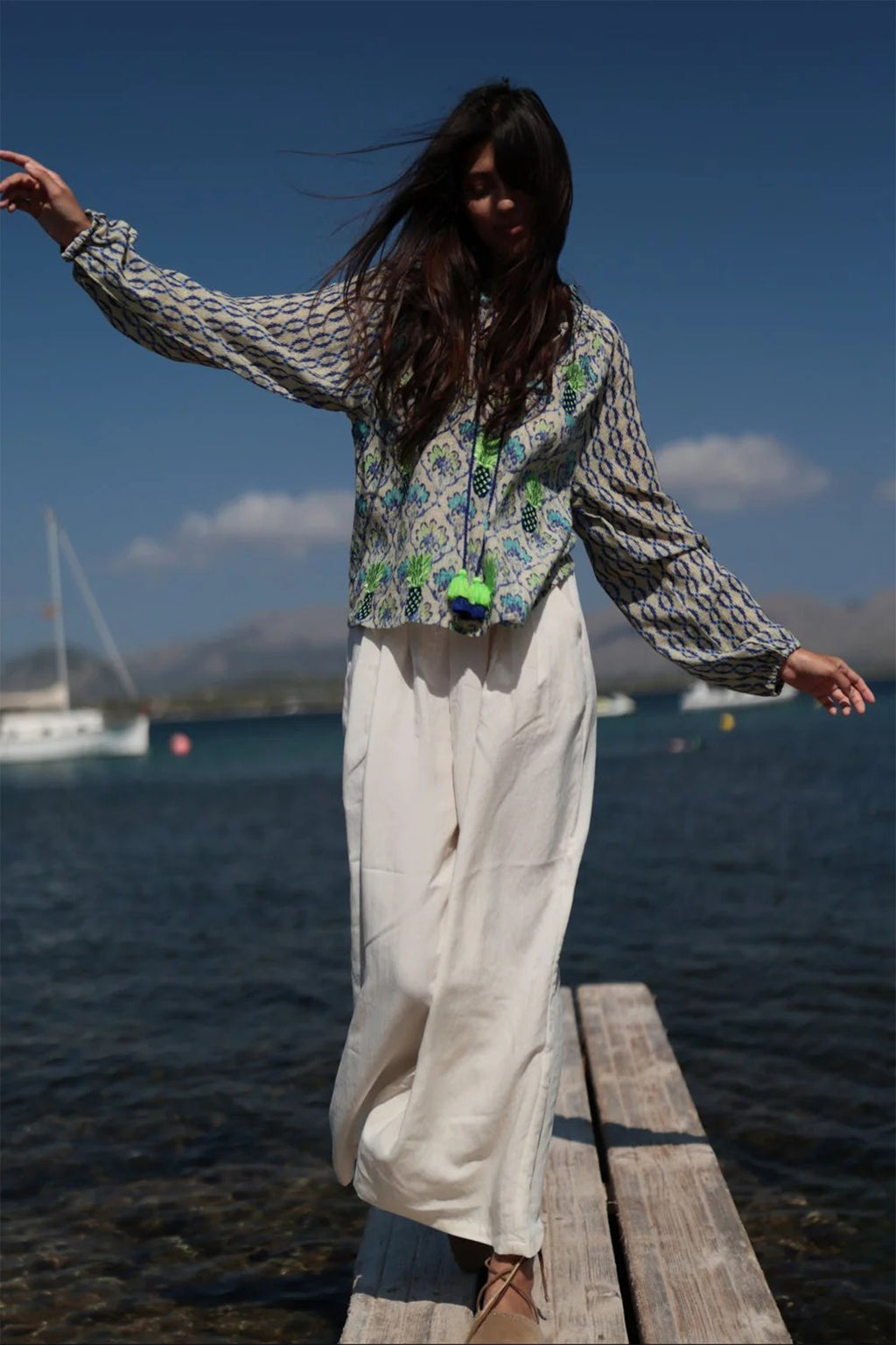 Woman standing on a dock by a body of water wearing a patterned top and white pants.